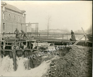 Photo d'une écluse dont les portes secouées par les dynamitages laisse fuir l'eau
    du bief amont tandis que deux hommes tentent de colmater les brèches.