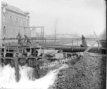 Photo d'une écluse dont les portes secouées par les dynamitages  laisse fuir l'eau du bief amont tandis que deux hommes tentent de colmater les brèches.